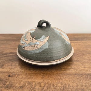 May include: A ceramic butter dish with a lid. The dish is dark green with a beige and blue wave pattern on the lid. The lid has a small, rounded handle. The dish sits on a wooden surface.
