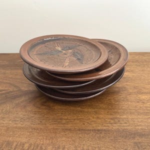 May include: A stack of four round ceramic plates with a brown glazed finish. The top plate features a dark, abstract, star-like pattern in the centre. Each plate has a raised rim and a smooth, glossy surface texture, showing earthy tones.