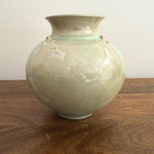May include: A round ceramic vase with a wide, flared rim and a smooth, glossy finish. The surface features a mottled blend of pale green, cream, and soft beige tones, creating a subtle, variegated appearance across the rounded body of the vessel.