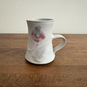 May include: A handmade ceramic mug with a white base and a curved handle. The mug features a floral design in red and blue. The mug is sitting on a wooden surface.
