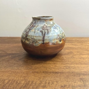 May include: A small, round ceramic vase with a wide base and a narrow opening. The vase has a brown base, a light blue and brown patterned middle section, and a darker brown rim. The vase is displayed on a wooden surface.