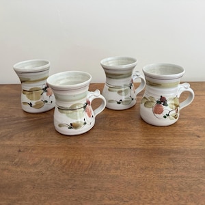 May include: A set of four ceramic mugs with a white base, featuring a flared rim and a tapered body. Each mug is decorated with hand-painted abstract brushstrokes in shades of sage green, brown, and muted orange, accented by thin black lines.