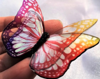 May include: A decorative butterfly ornament featuring translucent wings with a vibrant gradient of purple, pink, orange, and yellow. The wings display a white vein pattern and a subtle iridescent shimmer, connected by a textured black central body.