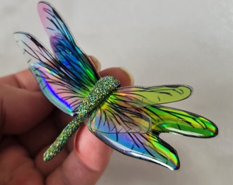 May include: A dragonfly ornament featuring iridescent, semi-transparent wings with black vein detailing. The body is textured with a dense, sparkling green glitter finish that catches the light. The wings display a vibrant spectrum of colors.