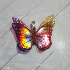 May include: A butterfly pendant featuring iridescent wings with a gradient of purple, yellow, and red hues. The wings display a detailed vein pattern and white spots along the edges. A metallic silver bail is attached to the center body.