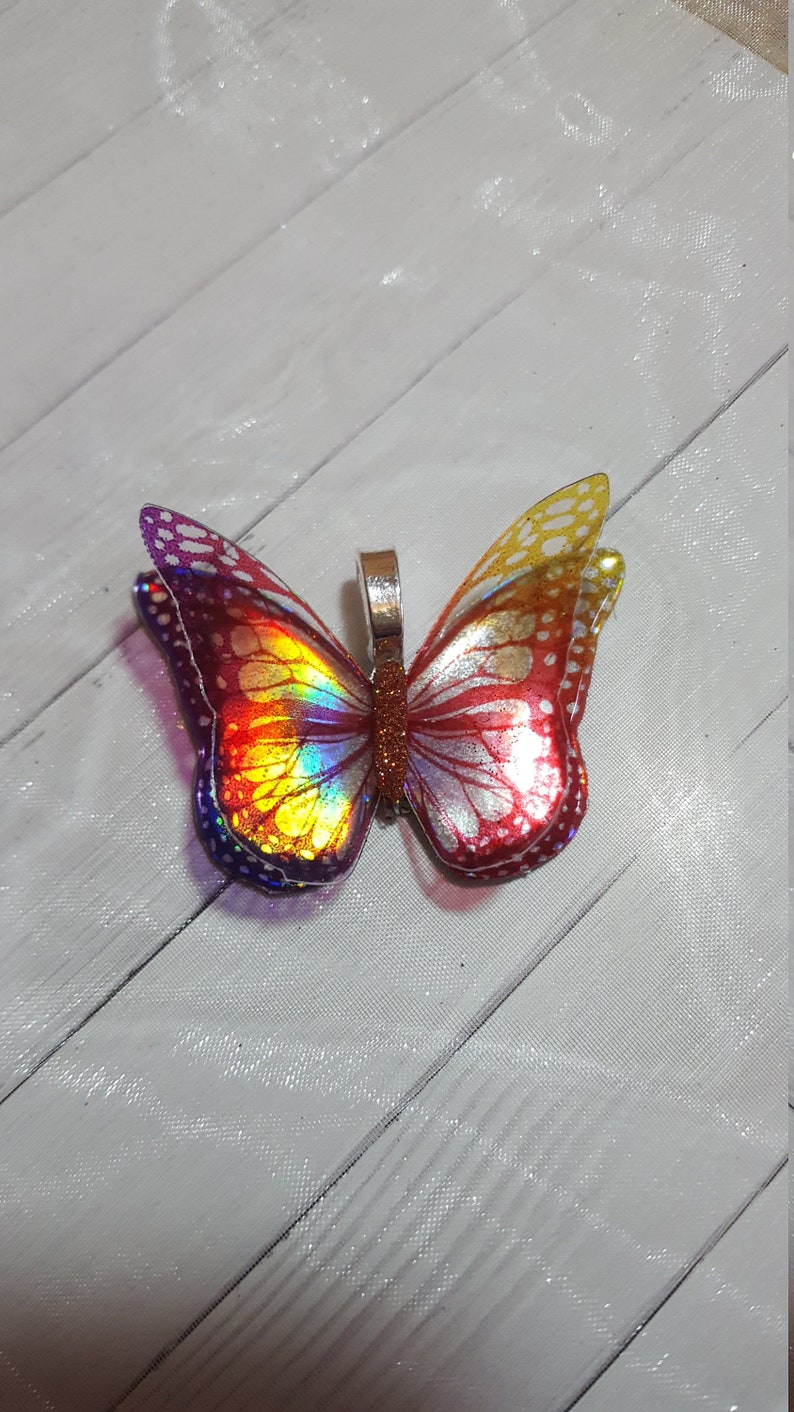 May include: A butterfly pendant featuring iridescent wings with a gradient of purple, yellow, and red hues. The wings display a detailed vein pattern and white spots along the edges. A metallic silver bail is attached to the center body.