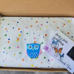 May include: A brown cardboard shipping box containing white tissue paper with a colorful polka dot pattern. A blue owl-shaped sticker with crystal accents is attached to the paper, alongside a white card featuring butterfly illustrations and text.