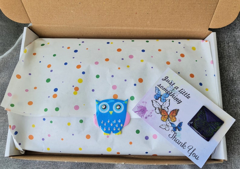 May include: A brown cardboard shipping box containing white tissue paper with a colorful polka dot pattern. A blue owl-shaped sticker with crystal accents is attached to the paper, alongside a white card featuring butterfly illustrations and text.