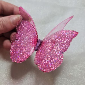 May include: A decorative butterfly ornament featuring translucent pink wings adorned with numerous sparkling pink rhinestones. The center body of the butterfly is covered in a textured purple glitter finish, creating a vibrant and reflective appearance.