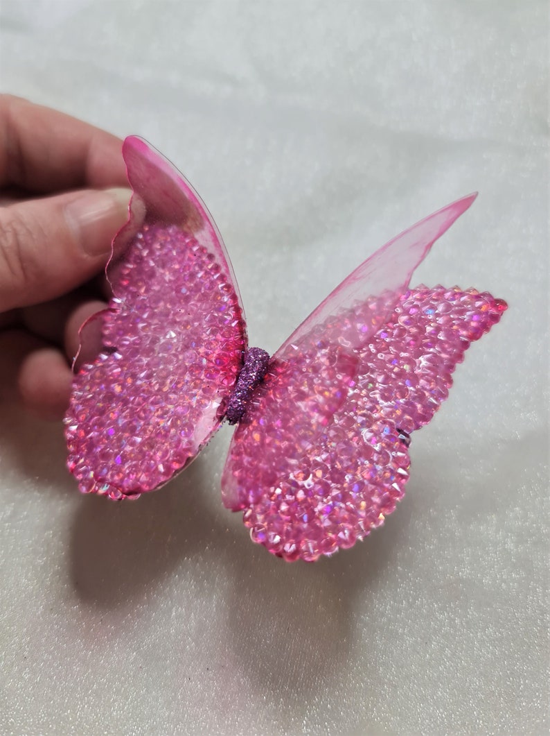 May include: A decorative butterfly ornament featuring translucent pink wings adorned with numerous sparkling pink rhinestones. The center body of the butterfly is covered in a textured purple glitter finish, creating a vibrant and reflective appearance.