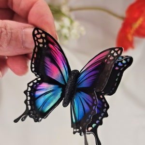 May include: A layered butterfly decoration featuring vibrant gradient wings in shades of pink, purple, and blue with black outlines and a textured black body. The wings have a glossy finish with iridescent accents along the edges and intricate cutouts.