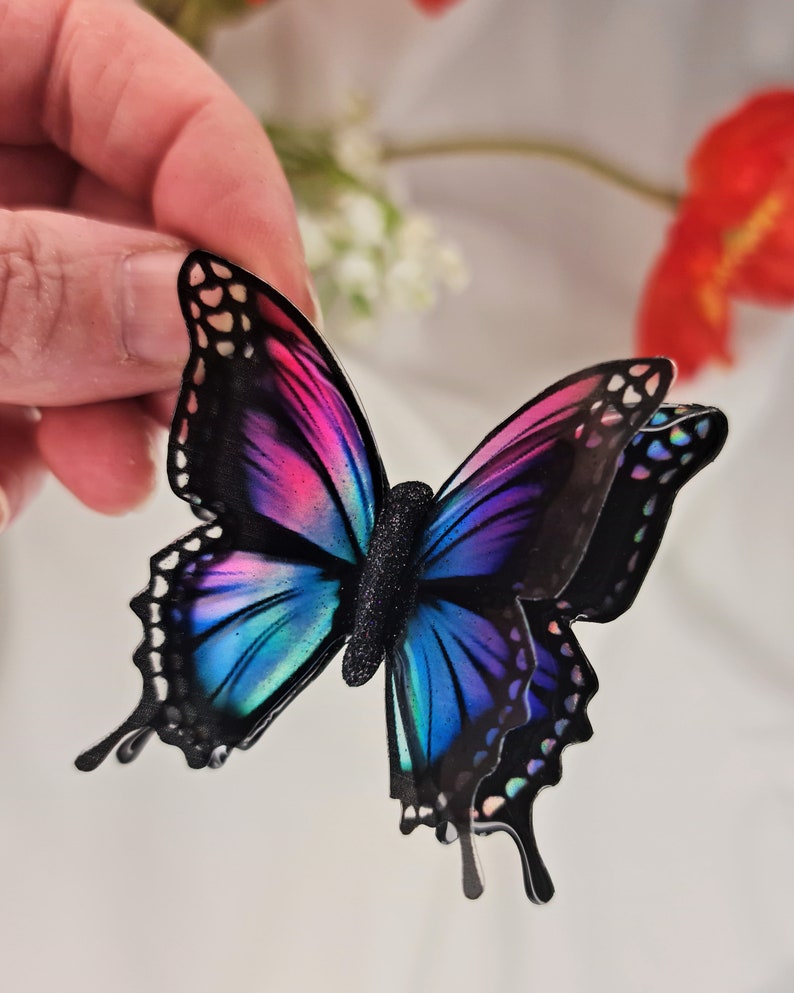 May include: A layered butterfly decoration featuring vibrant gradient wings in shades of pink, purple, and blue with black outlines and a textured black body. The wings have a glossy finish with iridescent accents along the edges and intricate cutouts.