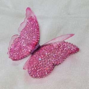 May include: A butterfly-shaped ornament featuring wings covered in small, sparkling pink crystals. The edges of the wings are made of a translucent, light pink material, and the central body is textured with a dark purple, glittery finish.