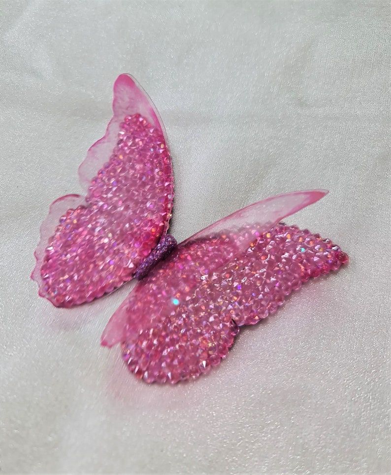 May include: A butterfly-shaped ornament featuring wings covered in small, sparkling pink crystals. The edges of the wings are made of a translucent, light pink material, and the central body is textured with a dark purple, glittery finish.