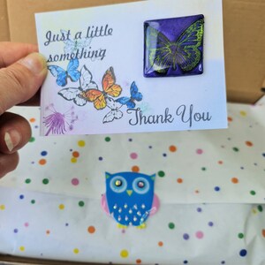 May include: A white card with the text Just a little something and Thank You, featuring printed butterflies and a square purple and green butterfly pin. Below, white tissue paper with colorful dots is sealed with a blue owl sticker.