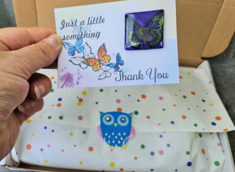 May include: A white card with the text Just a little something and Thank You, featuring printed butterflies and a square purple and green butterfly pin. Below, white tissue paper with colorful dots is sealed with a blue owl sticker.
