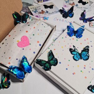 May include: Two white cardboard boxes decorated with colorful polka dots, containing white tissue paper and a pink heart cutout. Several iridescent blue and green butterfly ornaments are arranged on and around the boxes, with printed cards.
