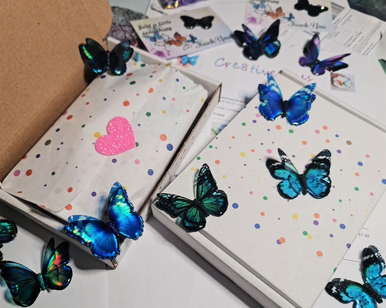May include: Two white cardboard boxes decorated with colorful polka dots, containing white tissue paper and a pink heart cutout. Several iridescent blue and green butterfly ornaments are arranged on and around the boxes, with printed cards.