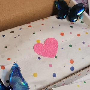 May include: A cardboard shipping box containing white tissue paper with a multicolored polka dot pattern. A bright pink glittery heart sticker is centered on the tissue paper. Two iridescent blue butterfly decorations are attached to the box.