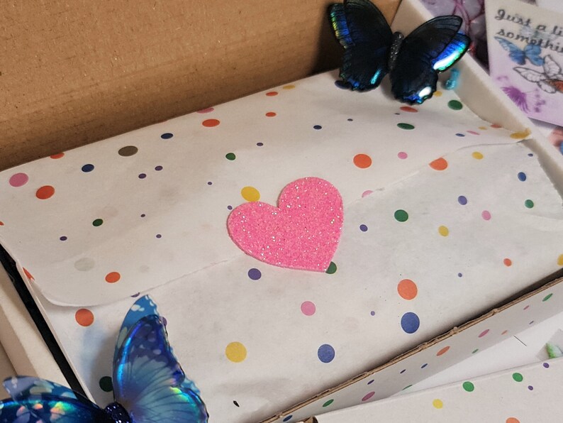 May include: A cardboard shipping box containing white tissue paper with a multicolored polka dot pattern. A bright pink glittery heart sticker is centered on the tissue paper. Two iridescent blue butterfly decorations are attached to the box.