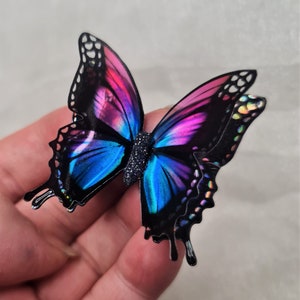 May include: A butterfly-shaped ornament featuring black wings with vibrant gradients of blue and magenta. The wings display iridescent, holographic-like spots along the edges, and the central body is textured with a dark, glittery finish.