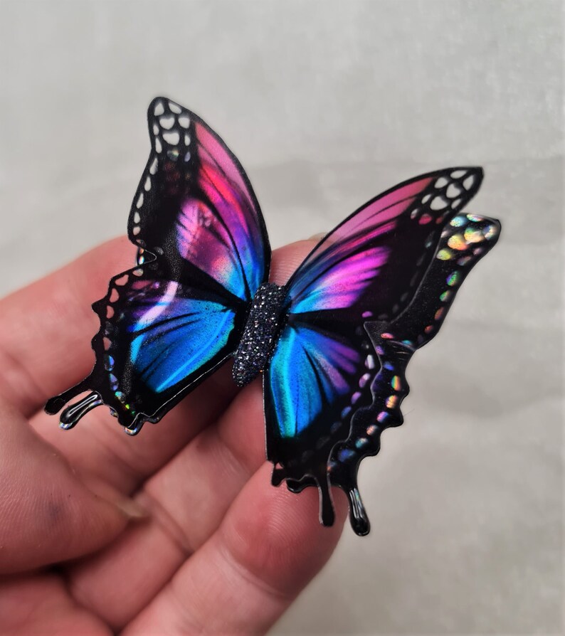 May include: A butterfly-shaped ornament featuring black wings with vibrant gradients of blue and magenta. The wings display iridescent, holographic-like spots along the edges, and the central body is textured with a dark, glittery finish.