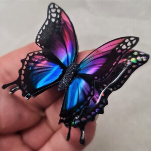 May include: A butterfly-shaped ornament featuring iridescent wings with a gradient of vibrant blue and magenta hues, outlined in black with white accents. The central body is textured with a dark, glittery finish, creating a reflective appearance.