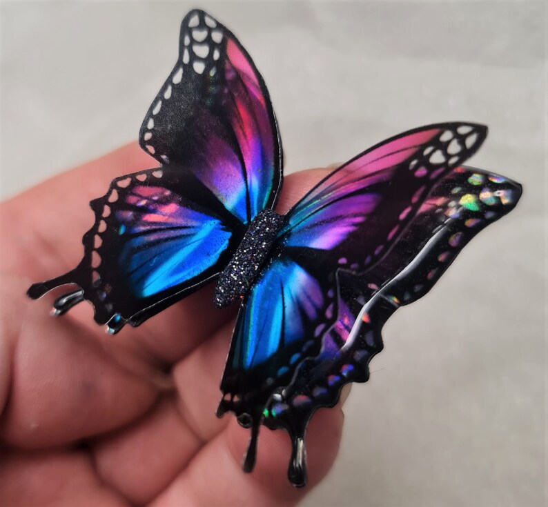 May include: A butterfly-shaped ornament featuring iridescent wings with a gradient of vibrant blue and magenta hues, outlined in black with white accents. The central body is textured with a dark, glittery finish, creating a reflective appearance.