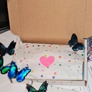 May include: A brown cardboard shipping box containing white tissue paper with multicolored polka dots, secured by a glittery pink heart sticker. Four iridescent blue and green butterfly decorations are placed around the box and tissue paper.
