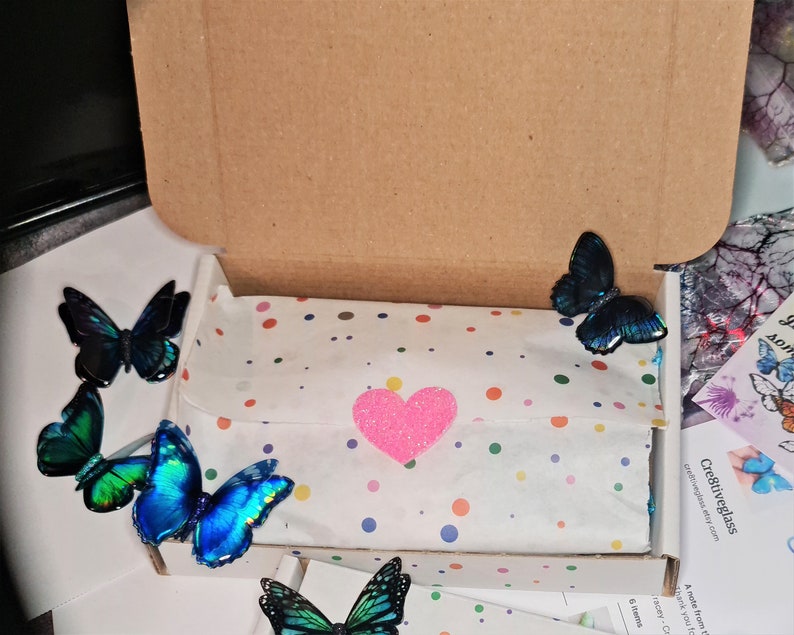 May include: A brown cardboard shipping box containing white tissue paper with multicolored polka dots, secured by a glittery pink heart sticker. Four iridescent blue and green butterfly decorations are placed around the box and tissue paper.