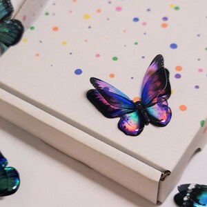 May include: A white cardboard box featuring a pattern of small, multicolored polka dots. Several decorative butterfly ornaments are arranged around the box, each displaying iridescent wings in shades of blue, purple, and green with black edges.