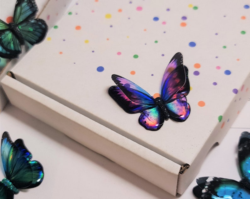 May include: A white cardboard box featuring a pattern of small, multicolored polka dots. Several decorative butterfly ornaments are arranged around the box, each displaying iridescent wings in shades of blue, purple, and green with black edges.