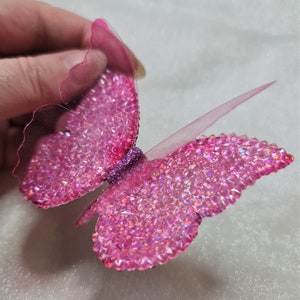 May include: A butterfly-shaped decorative ornament featuring translucent pink wings covered in small, sparkling pink rhinestones. The center of the butterfly is accented with a band of fine purple glitter, and the edges of the wings are clear.