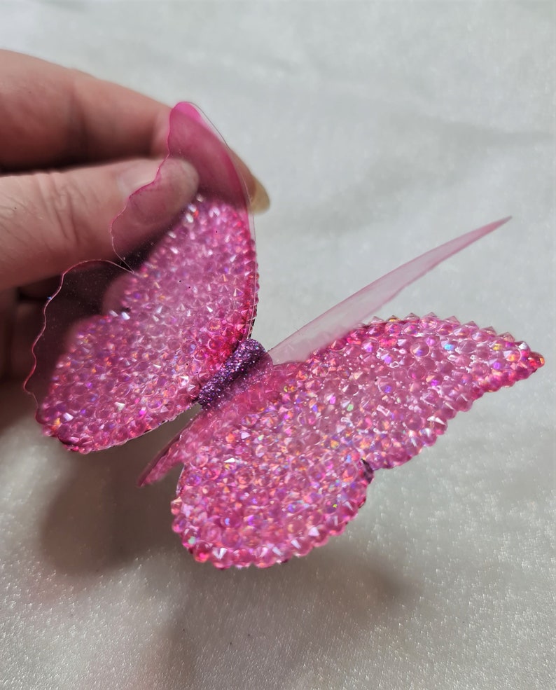 May include: A butterfly-shaped decorative ornament featuring translucent pink wings covered in small, sparkling pink rhinestones. The center of the butterfly is accented with a band of fine purple glitter, and the edges of the wings are clear.
