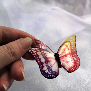 May include: A decorative butterfly ornament featuring translucent wings with a gradient color pattern transitioning from yellow and orange to red and deep purple, accented by a black textured body and defined black wing edges.