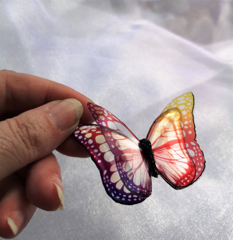 May include: A decorative butterfly ornament featuring translucent wings with a gradient color pattern transitioning from yellow and orange to red and deep purple, accented by a black textured body and defined black wing edges.