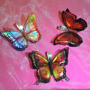 May include: Three butterfly-shaped pendants featuring iridescent wings. One pendant displays a holographic blue and orange pattern, another is deep red with a glittery center, and the third features a vibrant red, yellow, and black wing design.