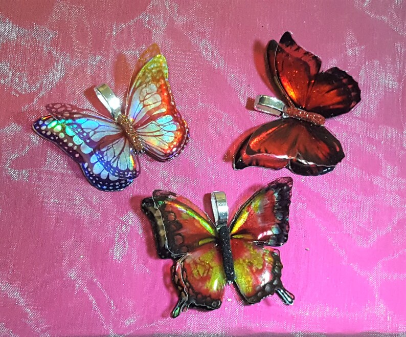 May include: Three butterfly-shaped pendants featuring iridescent wings. One pendant displays a holographic blue and orange pattern, another is deep red with a glittery center, and the third features a vibrant red, yellow, and black wing design.