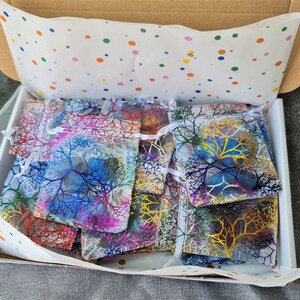 May include: A cardboard shipping box filled with multiple small, semi-transparent drawstring pouches. Each pouch features a colorful, branching, tree-like pattern in shades of blue, yellow, red, and purple. The box is lined with white tissue paper.