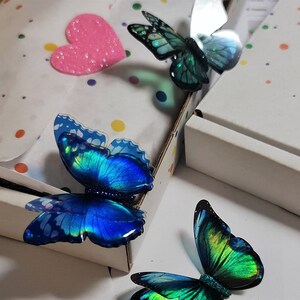 May include: Three decorative butterfly ornaments with iridescent blue and green wings featuring black edges and glittery bodies. The butterflies are arranged near a white cardboard box lined with white tissue paper decorated with colorful polka dots.
