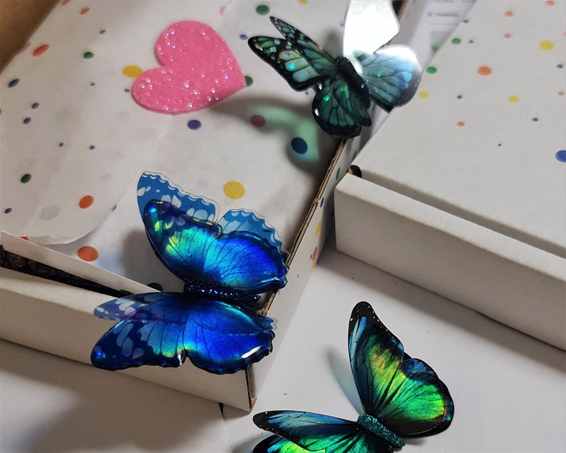 May include: Three decorative butterfly ornaments with iridescent blue and green wings featuring black edges and glittery bodies. The butterflies are arranged near a white cardboard box lined with white tissue paper decorated with colorful polka dots.