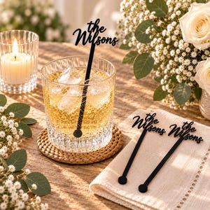 Könnte beinhalten: Drei schwarze Acryl-Getränkerührer mit dem personalisierten Text "the Wilsons" in einer kursiven Schriftart. Ein Rührer befindet sich in einem klaren Glas mit einer bernsteinfarbenen Flüssigkeit und Eiswürfeln, während zwei identische Rührer auf einer Serviette liegen.