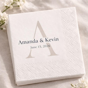 May include: A stack of white square napkins with a diamond pattern border. The napkins feature a large, light gray letter "A" and the names "Amanda & Kevin" with the date "June 15, 2026" printed in dark gray. Wedding theme.