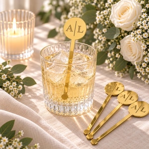 May include: A set of gold-toned metal drink stirrers featuring a circular top with the engraved letters A and L separated by a vertical line. One stirrer is placed inside a clear glass filled with ice and liquid, while three others lie nearby.