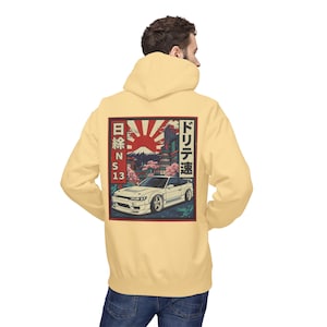 May include: A light yellow hoodie featuring a Japanese-inspired graphic on the back. The graphic includes a white car, Mount Fuji, a rising sun, and Japanese text. The hoodie is made of a soft material and has a hood and a front pocket.