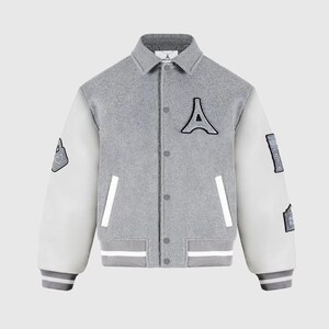 Op de afbeelding: Grijze varsity jas met witte mouwen. Voorzien van een grote zwarte "A" patch op de borst en kleinere patches op de mouwen. De jas heeft een knoopsluiting, witte zakafwerking en grijs-witte gestreepte manchetten en zoom.