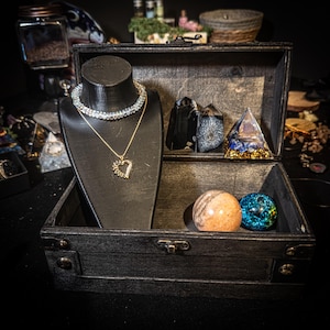 May include: A black wooden chest containing a black jewellery display bust with a crystal choker and a gold-tone heart pendant necklace. Inside the chest are two black crystal points, a blue and gold pyramid, and two spheres in tan and blue.