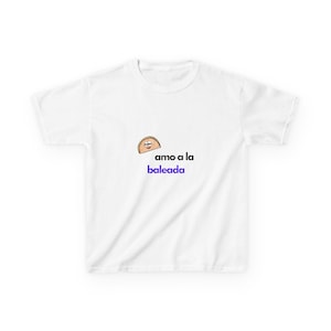 May include: White t-shirt featuring a cartoon baleada graphic with a smiling face. The text "amo a la baleada" is below the graphic in blue. A casual, comfortable tee.