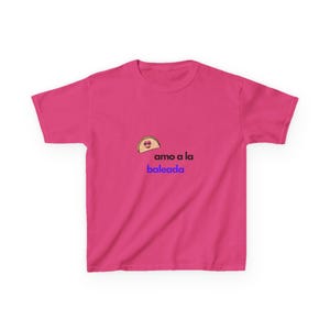 May include: A bright pink t-shirt featuring a cartoon baleada graphic with the text "amo a la baleada" in blue. The baleada graphic has a smiling face. The shirt is a casual wear item.