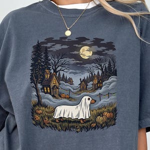 May include: A denim-colored t-shirt featuring a graphic print of a dog wearing a white ghost costume. The scene depicts a nighttime landscape with a full moon, dark trees, small houses, and pumpkins scattered across the grassy foreground.